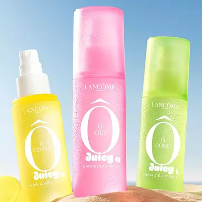 Dos frascos amarillos, uno de perfume rectangular con diseno texturizado y otro spray con tapa amarilla al costado, de pie sobre una playa de arena. Una toalla de playa rosada a rayas, doblada, se encuentra junto a los frascos. En el fondo, se aprecia un cielo azul despejado y el oceano.