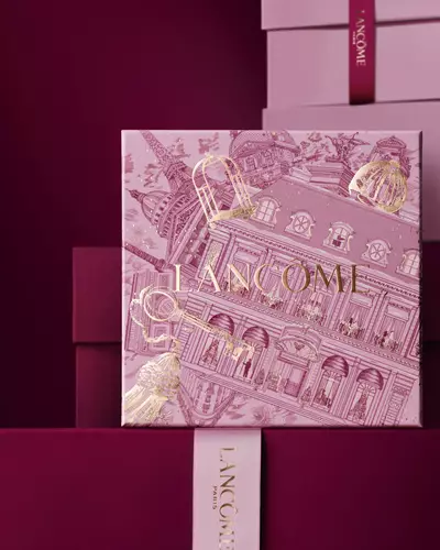 Imagen de una caja de regalo navidena rosa de Lancome frente a una pila de cajas de regalo rosadas y rojo oscuro, sobre un fondo rojo oscuro.