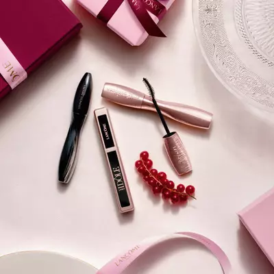 Una composicion vista desde arriba de productos de maquillaje Lancome, con mascara de pestanas y brillo labial, junto a cajas de regalo rosadas, cintas de saten y una ramita de grosellas rojas.