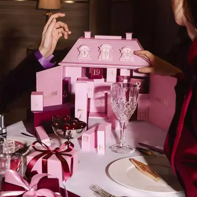 Manos interactuando con un calendario de adviento Lancome rosado sobre una mesa de comedor festiva con vajilla y cerezas.