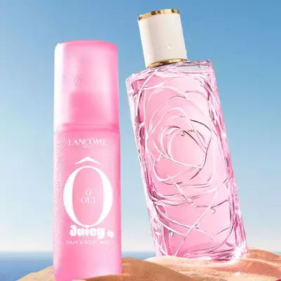Una escena de playa con tres coloridos frascos de perfume de pie sobre la arena, uno rosado, uno amarillo y uno verde, y otro frasco de perfume rosado acostado de lado en primer plano. A la izquierda, una toalla a rayas rosadas y blancas esta extendida sobre la arena. El fondo muestra un cielo azul despejado que se funde con un oceano azul en el horizonte.