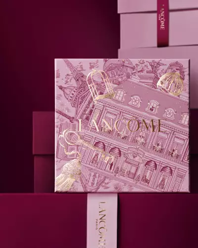 Imagen de una caja de regalo de fiestas rosada de Lancome frente a una pila de cajas de regalo rosadas y rojo oscuro, sobre un fondo rojo oscuro.